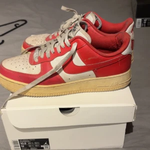 Nike Air Force 1 Low röd/vit - Nike Air Force 1 Low i rött och vitt med klassisk swoosh på sidorna och broderad Nike-logga på hälen. Skorna har snörning, platt sula och rund tå. Ovandelen är i skinn och textil, med vintage-inspirerad gulnad sula. Perfekt för dig som gillar retrostil.