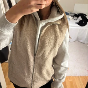 Beige fleeceväst med dragkedja - Mysig beige fleeceväst med hög krage och dragkedja framtill. Västen har två fickor på sidorna och är perfekt att slänga över en hoodie för en chill look. Den är enkel och stilren utan mönster, och har en mjuk teddystruktur.