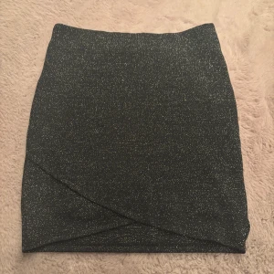 Glittrig svart kortkjol från Gina Tricot - Snygg svart kortkjol från Gina Tricot med glittriga inslag i tyget. Kjolen har omlott-look framtill och en tight passform som framhäver siluetten. Perfekt för dig som vill sticka ut med lite extra glitter.