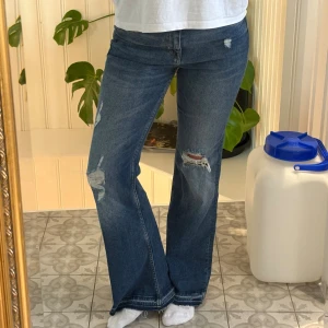 Blå bootcut jeans med slitningar - Säljer ett par blå jeans med bootcut-modell och snygga slitningar på benen. Jeansen har klassisk femficksdesign och är tillverkade i jeansmaterial. Perfekta för dig som gillar en avslappnad och trendig look.