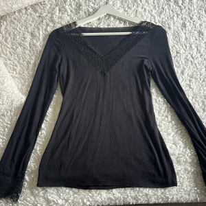Svart långärmad topp med spetsdetaljer - Svart långärmad topp från Intimissimi med snygga spetsdetaljer längs v-ringningen och vid ärmsluten. Toppens material är mjukt och stretchigt, vilket gör den bekväm och smickrande. Perfekt för dig som gillar en stilren look med en touch av spets.