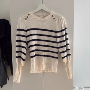  tröja från Neo Noir - Superfin tröja från Neo Noir med svarta ränder och snygga knappar på axlarna. Tröjan har puffiga långärmar och en klassisk rund halsringning. Perfekt för dig som gillar en stilren men trendig look.