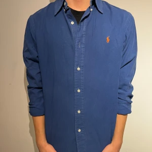 Mörkblå skjorta från Ralph Lauren S - Klassisk mörkblå skjorta från Ralph Lauren i bomull med custom fit. Skjortan har långa ärmar, knappar framtill och den ikoniska broderade loggan i orange på bröstet. Perfekt för en clean och stilren look.