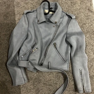 Ljusgrå bikerjacka från Zara Basic XS - Snygg ljusgrå bikerjacka från Zara Basic i mockaliknande material. Jackan har klassisk krage, axelklaffar, flera silvriga dragkedjor och bälte i midjan. Perfekt för dig som vill ha en edgy look med stilrena detaljer.
