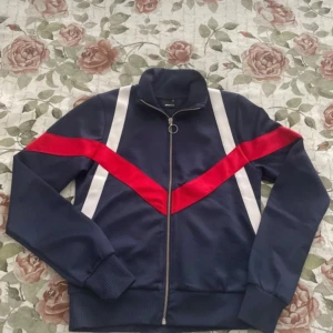 Blå ziptröja med röd och vit detalj Gina Tricot - Sportig blå ziptröja från Gina Tricot med vita och röda ränder i V-form både fram och bak. Tröjan har hög krage, långa ärmar och en snygg dragkedja med ring. Perfekt för en cool och avslappnad stil.