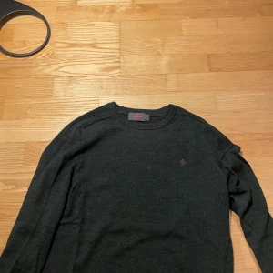 Mörkgrön merinoullströja från Morris - Säljer en stilren mörkgrön stickad tröja från Morris i 100% merinoull. Tröjan har rund halsringning, diskret broderad logga på bröstet och långa ärmar. Perfekt för dig som gillar klassisk och enkel stil.