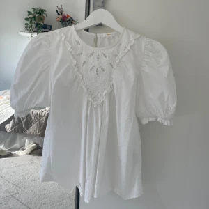 Vit blus med puffärm från Gina Tricot - Supersöt vit blus från Gina Tricot med korta puffärmar och broderad spetsdetalj framtill. Blusen har rund halsringning, volangkant och knäppning bak i nacken. Blusen är bara använd en gång, mycket bra skick.