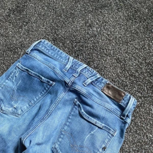 Replay Anbass blå slitna jeans - Säljer ett par blå Replay Anbass jeans med snygga slitningar och trasiga detaljer framtill. Jeansen har klassisk femficksdesign, normal passform och raka ben. Perfekta för dig som gillar en avslappnad och trendig look.