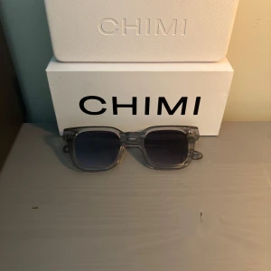 Grå transparenta solglasögon CHIMI - Snygga solglasögon från CHIMI med transparent grå båge och fyrkantig form. Glasen är mörka med en lätt blå ton. Kommer med originalväska och fodral. Perfekt accessoar för att lyfta din stil. Priset är justerbart!