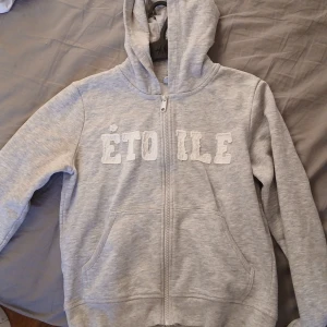 Grå zip up hoodie med text "☆ETOILE"  - Gråmelerad hoodie från Lindex med vit text 'ÉTOILE' framtill. Tröjan har dragkedja, huva och två fickor framtill. Mjuk insida och klassisk passform, perfekt för chill dagar. Storlek 170. Köpte för 229:-💓