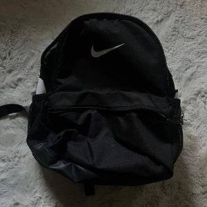 Liten Nike ryggsäck  - Svart liten ryggsäck från Nike. Väskan har ett stort huvudfack, ytterficka med dragkedja och meshfickor på båda sidorna. Justerbara, vadderade axelremmar. 