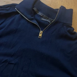 Mörkblå half zip tröja Jack & Jones - Mörkblå tröja från Jack & Jones med hög krage och snygg silverfärgad half zip-dragkedja. Tröjan är i mjukt material och har en klassisk, stilren look som passar perfekt till jeans eller chinos.
