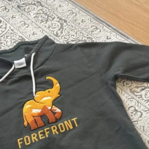 Mörkgrå hoodie med elefanttryck - Snygg mörkgrå hoodie från Forefront med broderad elefant i gult och orange på bröstet. Tröjan har huva med vita snören och lång ärm. Perfekt för dig som gillar unika detaljer och bekväma plagg.