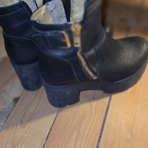 Svarta boots från Johnny Bulls - Säljer ett par svarta boots från Johnny Bulls med grov klack, dragkedja i guld och spänne vid skaftet. Skorna är i skinn och har en robust, chunky sula som ger en cool look. Perfekta för dig som gillar en tuffare stil.