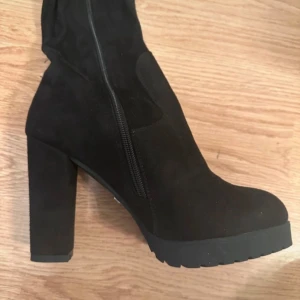 Svarta stövlar med klack från Tiamo - Snygga svarta stövlar från Tiamo med hög blockklack och platåsula. Skorna har dragkedja på sidan för enkel påtagning och en stilren, rund tå. Perfekta för att lyfta din outfit med en edgy vibe