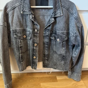 Grå croppad jeansjacka från &Denim - Snygg croppad jeansjacka från &Denim i tvättad grå färg. Jackan har rå, fransig nederkant, klassisk krage, fejk bröstfickor med knappar och långa ärmar. Perfekt för dig som gillar en avslappnad och trendig stil.💕 Säljer då den inte längre kommer till användning