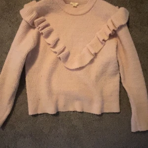 Ljusrosa stickad tröja med volang - Superfin ljusrosa stickad tröja med lång ärm och snygg volangdetalj som går diagonalt över bröstet och axeln. Tröjan har ett diskret vågmönster i stickningen och rund hals. Perfekt för dig som gillar gulliga detaljer och mjuka pasteller.