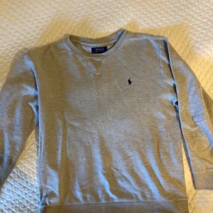 Grå sweatshirt från Polo Ralph Lauren - Säljer en stilren grå sweatshirt från Polo Ralph Lauren med klassisk rund halsringning och broderad logga på bröstet. Tröjan är långärmad och gjord i mjuk bomull, perfekt för chill dagar eller när du vill ha en clean look. Väldigt lite använd. Passar  XS S man o även kille 14-16 år