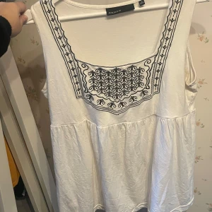 Vit ärmlös blus från KappAhl - Supersöt vit blus från KappAhl i storlek M. Den är ärmlös och har ett broderat svart mönster framtill vid halsringningen. Blusen har en lös passform och är tillverkad i mjuk bomull, perfekt för varma dagar.