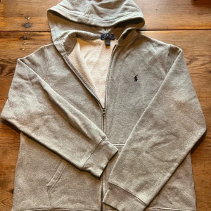 Grå hoodie från Polo Ralph Lauren XL - Grå zip-hoodie från Polo Ralph Lauren i storlek XL, men passar även M. Knappt använd och i mycket fint skick. Klassisk modell med dragkedja, huva, fickor och broderad Polo-logga. Skriv vid frågor – priset är inte hugget i sten.