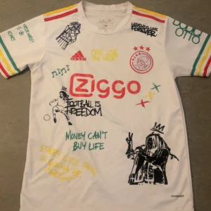 Ajax x Bob Marley vit fotbollströja - Unik Ajax fotbollströja i vitt från Adidas med färgglada detaljer på ärmarna, tryckta Bob Marley-citat och illustrationer. Ziggo-logga på bröstet, Ajax-emblem och coola grafiska prints över hela tröjan. Materialet är lätt och andas, perfekt för fotboll eller streetwear.