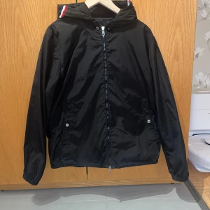 Svart vindjacka från Moncler - Svart vindjacka från Moncler med huva och dragkedja framtill. Jackan har Moncler-logga på ärmen och rödvit rand på huvan. Två fickor med tryckknappar och elastiska muddar. Perfekt för dig som vill ha en stilren och sportig look. Storlek m/s