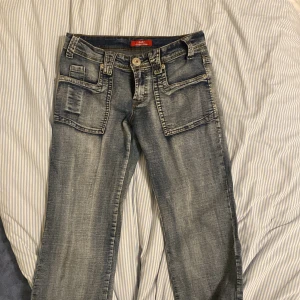 Blå jeans med stora fickor G-STAR RAW - Säljer ett par blå jeans från G-STAR RAW med stora fyrkantiga fickor fram och bak. Jeansen har en snygg tvättad look, raka ben och klassisk knappgylf. Perfekta för dig som gillar streetstyle och vill sticka ut lite extra.