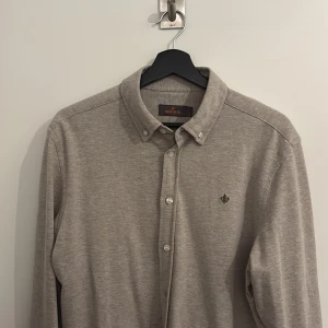 Beige skjorta från Morris Stockholm - Snygg beige skjorta från Morris Stockholm med klassisk krage och knappar framtill. Skjortan har lång ärm och en liten broderad logga på bröstet. Perfekt för dig som gillar stilrena och enkla plagg med en exklusiv touch.