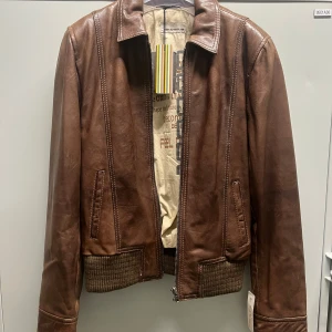 Brun skinnjacka Rock'n Blue S - Snygg brun skinnjacka från Rock'n Blue i modell Ziggy Spring. Jackan har ribbade muddar, dragkedja framtill och klassisk krage. Insidan är fodrad med beige tyg och har flera innerfickor. Perfekt för dig som vill ha en tidlös och cool look.