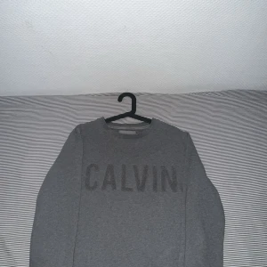 Grå Calvin Klein Jeans tröja - Snygg grå tröja från Calvin Klein Jeans med stort 'CALVIN' tryck framtill. Klassisk rund halsringning och långa ärmar. Mjuk och skön bomullsblandning, perfekt för en clean och stilren look.