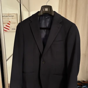 Mörkblå kostym från Riley  - Snygg mörkblå kostym från Riley med klassisk kavaj och matchande kostymbyxor. Kavajen har två knappar, smala slag och flera fickor. Storlek på kavaj är 48 och byxorna 50. Nypris cirka 2000kr