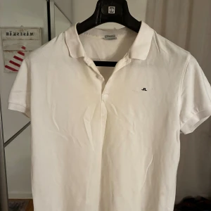 Vit pikétröja från J.Lindeberg - Klassisk vit pikétröja från J.Lindeberg med liten broderad logga på bröstet. Tröjan har korta ärmar, krage och knäppning framtill. Perfekt för en clean och sportig look. Nypris cirka 400kr.