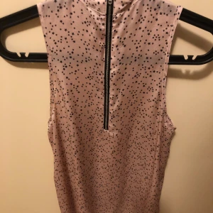 Rosa ärmlös topp med stjärnmönster - Säljer en ljusrosa ärmlös topp med svarta små stjärnor över hela tyget. Toppen har en synlig dragkedja i ryggen som ger en cool detalj. Perfekt att matcha med jeans eller kjol för en trendig look.