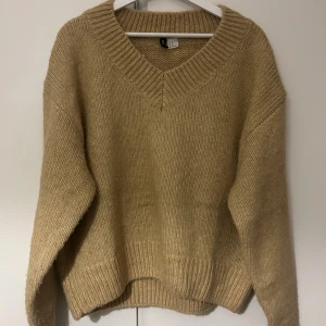 Beige stickad v-ringad tröja Divided - Mysig beige stickad tröja från Divided med v-ringad hals och bred ribbad kant. Tröjan har en loose passform och långa ärmar, perfekt för lager på lager under höst och vinter. Enkel att matcha med jeans eller kjol.