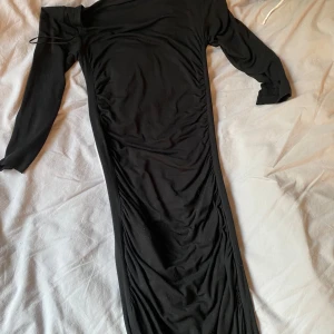 Svart långklänning från H&M Basic - Svart långklänning från H&M Basic med figurnära passform och trekvartsärm. Klänningen har lätt rynkade detaljer längs sidorna och en enkel, stilren design. Perfekt för dig som gillar minimalistisk stil och vill ha en klänning som känns både bekväm och snygg.