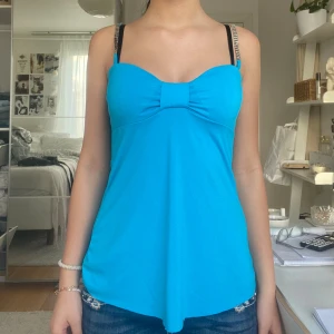 Turkos linnetopp med strassband - Supersöt turkos linnetopp med smala axelband dekorerade med strass. Toppen har en söt rosettdetalj framtill och en lätt utsvängd passform. Perfekt för dig som vill sticka ut med färg och bling. Materialet känns mjukt och stretchigt.