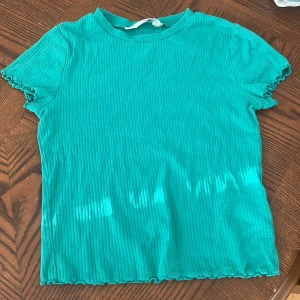 Turkos ribbad topp från H&M - Säljer en turkos ribbad topp från H&M med korta ärmar och lätt croppad passform. Toppen har en rund halsringning och vågiga kanter vid ärmslut och nederkant. Perfekt för dig som gillar färg och enkel stil.