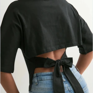 Nelly topp - Svart topp i bomull med halvlånga ärmar och rund hals. Baksidan har en öppen rygg med breda band som knyts i en stor rosett. Snygg, figurnära passform som markerar midjan och ger en modern look. Har används 1-2 gånger