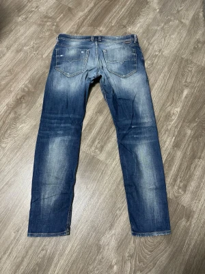 Blå slitna jeans med raka ben - Snygga blå jeans med slitningar och ljusa partier framtill. Jeansen har raka ben, klassiska fem fickor och en cool, lite sliten look. Perfekta för dig som gillar en avslappnad och trendig stil.
