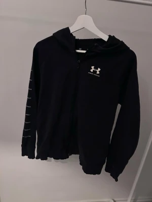 Svart hoodie från Under Armour - Säljer en svart hoodie från Under Armour med dragkedja framtill och vit logga på bröstet. Hoodien har huva och diskreta detaljer på ena ärmen. Tillverkad i mjukt och bekvämt material, perfekt för en sportig och avslappnad stil.