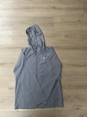 Grå vindjacka från Under Armour - Snygg grå vindjacka från Under Armour med huva och dragkedja framtill. Jackan har logga på bröstet och stor text på ryggen. Tillverkad i lätt syntetmaterial, perfekt för blåsiga dagar. Två sidofickor och långärmad modell. Jackan är i storlek 2Xl men sitter som Xl