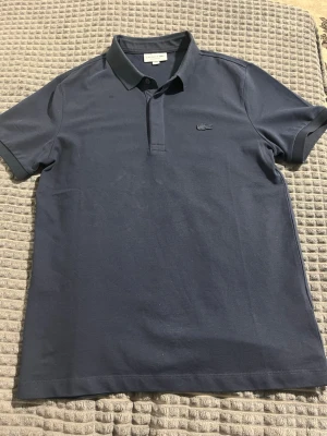 Mörkblå pikétröja från Lacoste - Klassisk mörkblå pikétröja från Lacoste med korta ärmar och krage. Tröjan har den ikoniska krokodilloggan broderad på bröstet och är tillverkad i bomull för en bekväm känsla.Även så sitter den tajt mellan bicepsen och brösten som får dig och se ut väll tränad, ny pris på den ligger mellan 900krup till 1400kr 