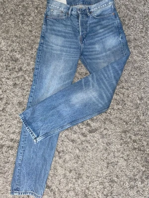 Blå raka jeans från H&M - Klassiska blå jeans från H&M med rak passform och fem fickor. Jeansen har en ljus tvätt och subtila slitningar för en avslappnad look. Perfekta till vardags och lätta att matcha med olika stilar.