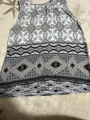 Mönstrad ärmlös topp Soyaconcept - Snygg ärmlös topp från Soyaconcept med detaljerat grafiskt mönster i svart, grått och beige på vit botten. Lätt och luftig modell med rund halsringning, perfekt för en trendig och avslappnad look.