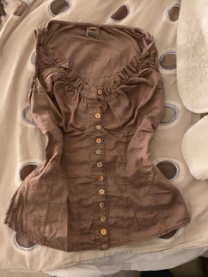 Brun korsettopp med knappar - Säljer en brun korsettopp från Community Co. med snygg v-ringning och rynkade detaljer vid bysten. Toppen har dekorativa knappar hela vägen framtill och är figurnära i modellen. Perfekt att styla med jeans eller kjol för en trendig look.