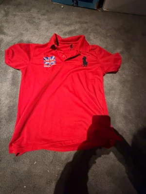 Röd pikétröja från Polo Ralph Lauren - Snygg röd pikétröja från Polo Ralph Lauren med klassisk krage och knappar. Tröjan har en stor svart logga på bröstet och en engelsk flagga med texten 'England' på andra sidan. Perfekt för dig som gillar sportig och stilren look.