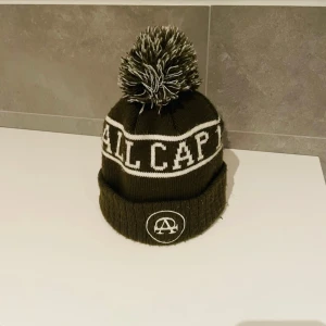 Allcap 1 mössa - Säljer en mörkgrön stickad mössa från All Cap med vit text och logga framtill. Mössan har en stor tofs i svart och vitt samt vikt kant. Perfekt för kalla dagar och ger en cool streetwear-look.