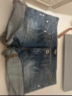Blå jeansshorts med uppvik - Säljer ett par blå jeansshorts från Heart Quake med klassisk femficksdesign och uppvikta benslut. Shortsen har en normal passform och detaljer i silverfärgade nitar. Perfekta för sommaren och lätta att matcha med olika stilar.
