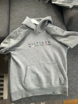 Grå hoodie från Tommy Hilfiger - Snygg grå hoodie från Tommy Hilfiger med klassisk logga och texten 'Hilfiger New York' på bröstet. Hoodien har en stor känguruficka framtill, huva och små broderade detaljer på ärmen. Tillverkad i mjukt sweatshirtmaterial, perfekt för en chill och avslappnad stil.