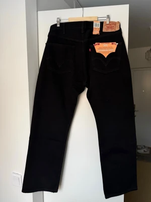 Levi's 501 svarta jeans straight fit - Klassiska svarta Levi's 501 jeans med rak passform och femficksdesign. Jeansen har knappgylf och är tillverkade i slitstark bomull. Perfekta för dig som gillar tidlös stil och vill ha ett par riktigt snygga jeans i garderoben. Översta knappen är trasig och behöver bytas ut.